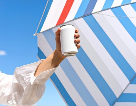 Brazo sosteniendo una lata o botella blanca en alto, con un paraguas de playa de rayas azules y rojas y cielo azul claro de fondo