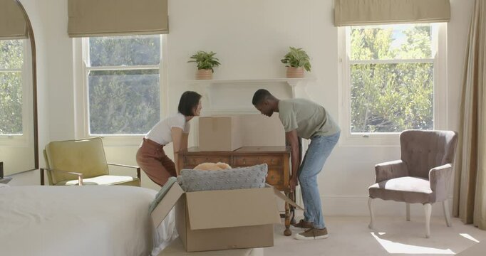 Diverse couple exchanging look lifting placing desk to set up bedroom unpacking pillows from boxes