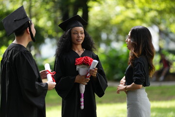 Three graduates in caps and gowns celebrating outdoors. One holds a diploma and bouquet, while the...