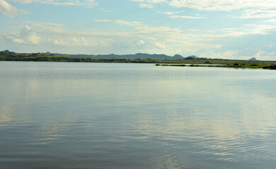 The flat surface of a large lake surrounded by high mountains covered with pine forests under a cloudy summer sky.