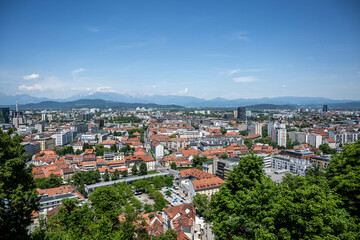 Fototapeta premium Panoramic view of Ljubljana city center featuring traditional architecture with terracotta roofs, modern buildings, and Julian Alps backdrop under clear blue sky