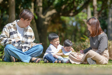 Fototapeta premium In March, at a sunny forested park in Japan, a Japanese couple sits on the grass with their 3-year-old son and 6-month-old baby between them. This photo shows a quiet and loving spring family moment.