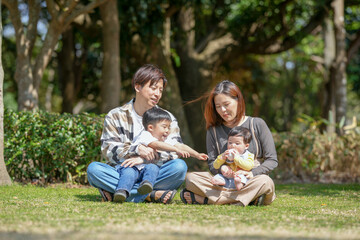 Fototapeta premium In March, at a sunny forested park in Japan, a Japanese couple sits on the grass with their 3-year-old son and 6-month-old baby between them. This photo shows a quiet and loving spring family moment.
