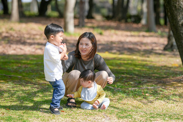 In March, at a sunny forested park in Japan, a Japanese mom and her 6-month-old baby sit on the grass. Nearby, her 3-year-old son tosses a ball. This photo captures a peaceful spring family moment.