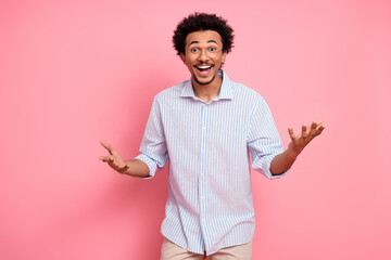 Stylish young man with curly hair making a cheerful gesture on a pink background in casual striped...