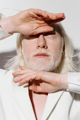 Artistic portrait of a person with white hair and beard, hands raised, against a minimalist light...