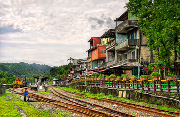 Colorful hillside houses overlook the curving railway tracks and a small diesel locomotive at Shifen Station on the Pingxi line in New Taipei, Taiwan, with lush forested hills beneath a cloudy summer