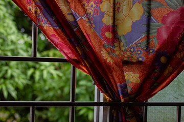 Colorful, vibrant patterned curtain draped by a metal window frame, with a blurred green garden...