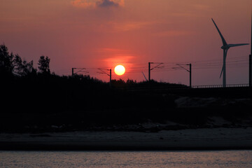 A stunning sunset paints the sky in shades of red and orange over Houlong Port, Miaoli County, Taiwan. A wind turbine and railway overhead lines are silhouetted against the bright sun.
