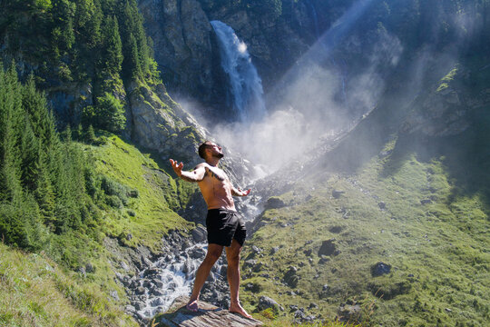 Carefree man in nature. Freedom man in alpine landscape. Muscular model breathing air near waterfall. Handsome model with raised hands outdoors. Fit lifestyle. Relaxed man. Sexy man. Healthy torso.