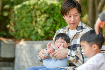 In a sunlit forest park in March, a 3-year-old Japanese boy shows a bird feather to his father, who is in his 30s and holding a six-month-old baby boy in his arms, sharing a quiet family moment.