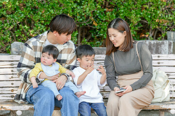 In March, at a sunny park in Fukuoka, a Japanese couple in their thirties sits on a bench with their 3-year-old son and baby girl, 6 months old. This photo captures a peaceful family moment outdoors.