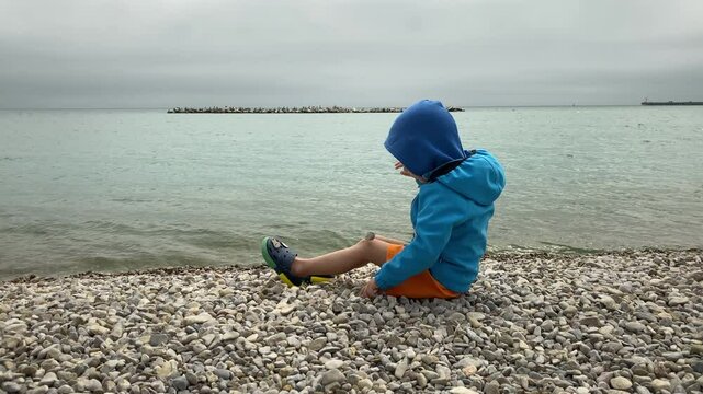 A young child, dressed in a blue hooded jacket and orange shorts with green crocs, sits quietly on a rocky pebble beach, gazing down towards the water