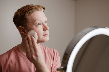A Close-up Of A Young Man Applying Foundation Skincare Makeup Routine
A young man applies foundation using a makeup sponge while standing in front of a mirror with an LED ring light. 