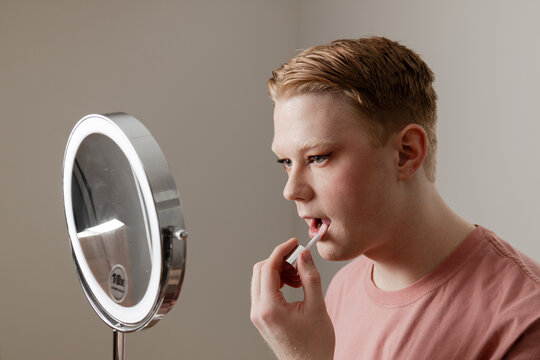 A Medium shot of a Man Applying Lip Gloss in Front of Mirror
A young man applies lip gloss while looking into a vanity mirror with LED lighting. Image highlights themes of self-expression, confidence. - Powered by Adobe