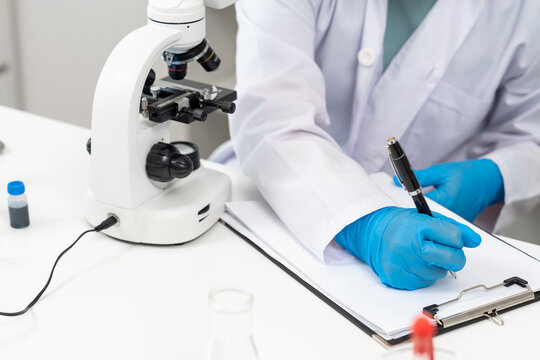 Close-up of hand wearing blue glove writing with pen on clipboard beside white microscope. Laboratory desk contains scientific tools including chemical vial, beaker and lab notebook used in experiment