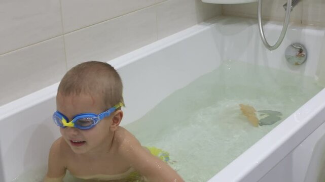 little boy with swimming goggles on is sitting in a white bathtub full of water. He is learning to hold his breath while diving underwater.
