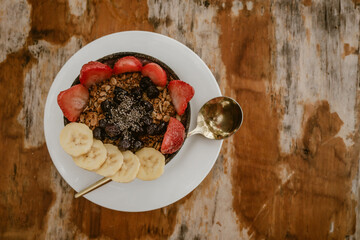 Delicious healthy smoothie bowl served in a coconut and topped with fresh strawberries, banana slices and dried berries on a white plate with golden spoon during a summer holiday in Asia.
