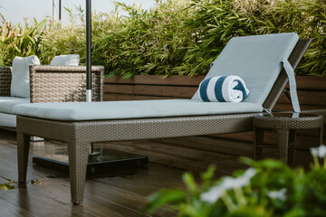 A lounge chair with a soft cushion and a blue and white rolled up towel surrounded by lush greenery outdoor at daytime along the pool of a hotel in Manila during a summer holiday in the Philippines.