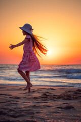 Ocean beach sunrise and happy little girl with hat and summer dress walking and dancing on the sandy shore, her silhouette is against the rising sun