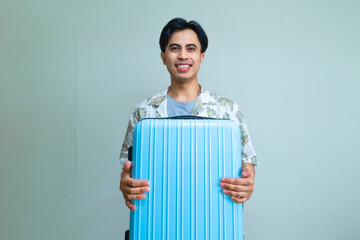 Smiling young Asian man in summer clothes carrying and hugging a suitcase, looking cheerful and ready for vacation, posing against a clean, colorful background for travel or holiday concepts