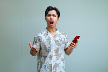 Angry young Asian man in summer clothes holding a smartphone and staring at the camera with a serious expression, isolated on a clean studio background