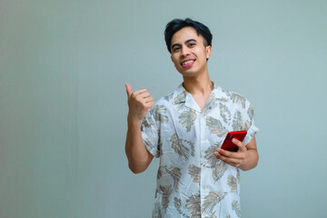 Happy young Asian man in summer clothes pointing aside with his thumb while holding a smartphone, isolated on a clear background, showing excitement and joy