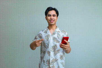 Portrait of a smiling young Asian man in summer clothes pointing at a cellphone he is holding, isolated on a clear background, perfect for digital ads, promotions, or mobile app concepts