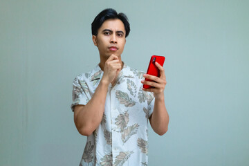 Serious young Asian man wearing casual summer clothes, holding a smartphone and touching chin with a thoughtful expression, isolated on a clean studio background