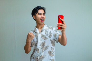 Happy young Asian man wearing summer clothes celebrates success with clenched fist while holding smartphone, excited and smiling on a clear background, ideal for promotions