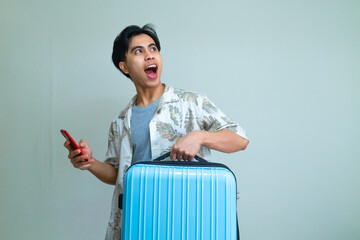 Happy young Asian man in summer clothes looking to the side with an excited expression, holding a smartphone and suitcase, standing against a clear background with copy space