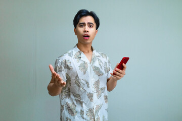 Shocked young Asian man in summer outfit holding a smartphone, reacting with surprise against a clear background, perfect for digital ads, mobile apps, or promotional content