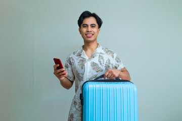 Happy young Asian man in summer outfit smiling while using smartphone and carrying a blue suitcase, ready for travel or vacation, isolated on a clear background
