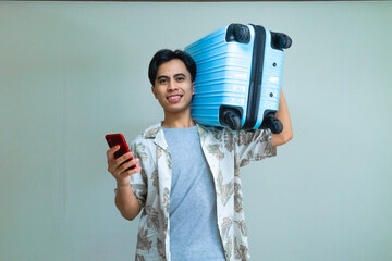 Happy young Asian man in summer outfit smiling while using smartphone and carrying a blue suitcase, ready for travel or vacation, isolated on a clear background