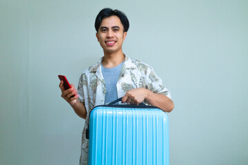 Happy young Asian man in summer outfit smiling while using smartphone and carrying a blue suitcase, ready for travel or vacation, isolated on a clear background