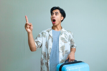 Portrait of a young Asian man in summer clothes pointing to the side with a funny wow expression while carrying a suitcase, isolated on a clear background, travel concept
