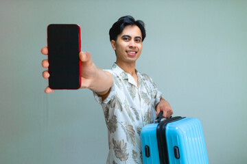 Smiling young Asian man in summer outfit showing blank mobile phone screen and carrying a blue suitcase, perfect for travel app, booking service, or holiday advertisement concept