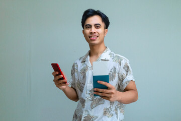 Happy young Asian man in summer outfit holding passport and plane ticket while using smartphone, excited for vacation or travel, isolated on a clear background