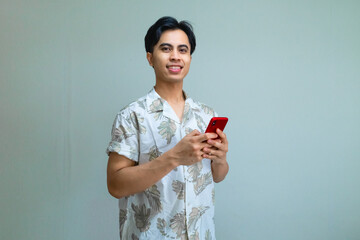 Portrait of a smiling young Asian man wearing summer clothes, confidently using a cell phone on a clean clear background, radiating a relaxed and positive mood