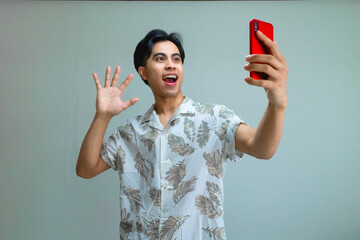 Happy young Asian man in summer outfit recording himself with a smartphone, smiling and making a fun gesture against a clean, clear background, perfect for social media or advertisement use