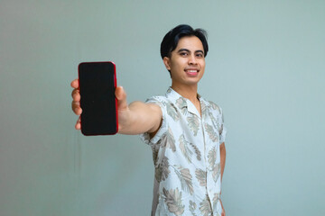 Happy young Asian man in summer outfit showing a blank mobile phone screen, standing against a clean background, perfect for advertising, app promo, or digital marketing