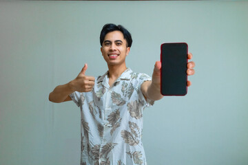 Happy young Asian man in summer outfit giving a thumbs-up while showing a blank mobile phone screen, standing against a clear background, perfect for app promo or advertisement