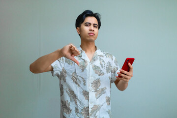 Serious young Asian man in summer clothes holding smartphone and showing thumbs down gesture, standing against a clear background, expressing disapproval or negative feedback