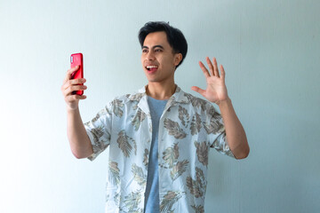Portrait of a smiling young Asian man in summer clothes making a video call gesture, isolated on a clear background, cheerful and friendly expression perfect for digital ads or promotions