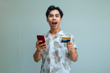 appy young Asian man in summer outfit holding a credit card and using a smartphone for online transaction, showing amazed face expression against a clear background