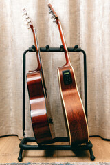 Two acoustic guitars stand side-by-side on a black metal guitar stand in front of beige curtains