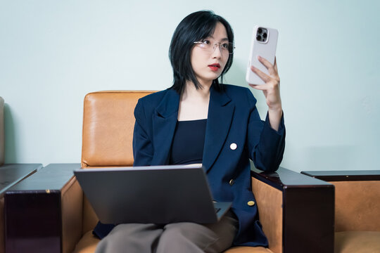 Woman in a modern lounge checking her smartphone while working on a laptop in a stylish indoor setting during daylight hours