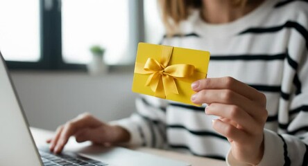 Woman typing on laptop keyboard while holding yellow gift card adorned with gold ribbon, representing online shopping convenience and digital transaction ease