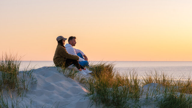 A couple sits on a sandy dune, surrounded by tall grass, admiring the vibrant sunset over the calm waters of the beach in Denmark. The warm hues create a romantic atmosphere. Vejers Strand beach