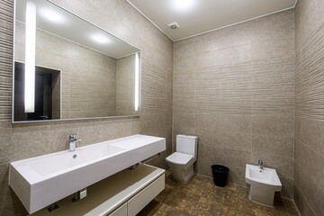 Modern bathroom with beige textured tiles, white fixtures (sink, toilet, bidet), a large mirror with lights, and dark brown floor tiles. Clean and contemporary style